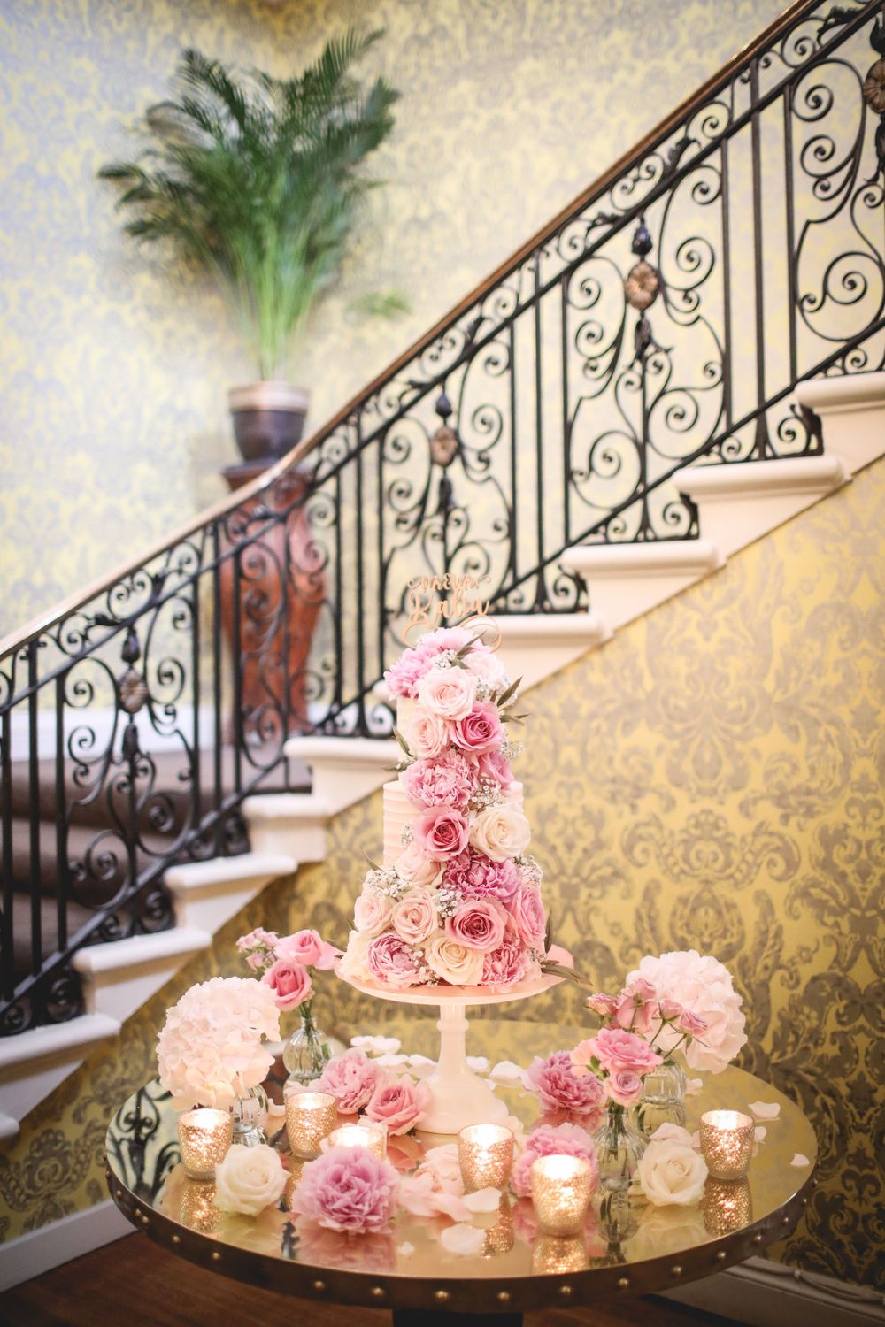 Elegant pink wedding cake displayed on a mirrored table near an ornate staircase with wrought iron railing. Hindu Wedding Photography