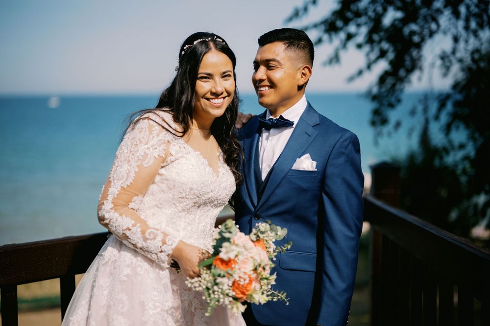 A couple in wedding attire smile by the ocean, with the bride in lace and groom in navy suit holding a peach bouquet.