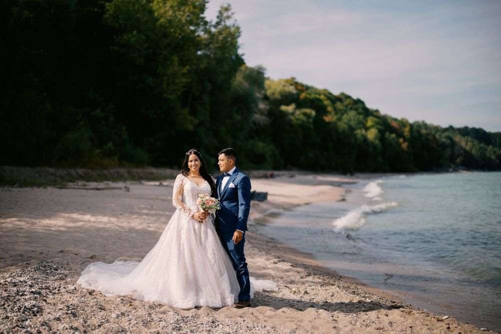 A wedding couple walks along a scenic beach shoreline with trees and water in the background.