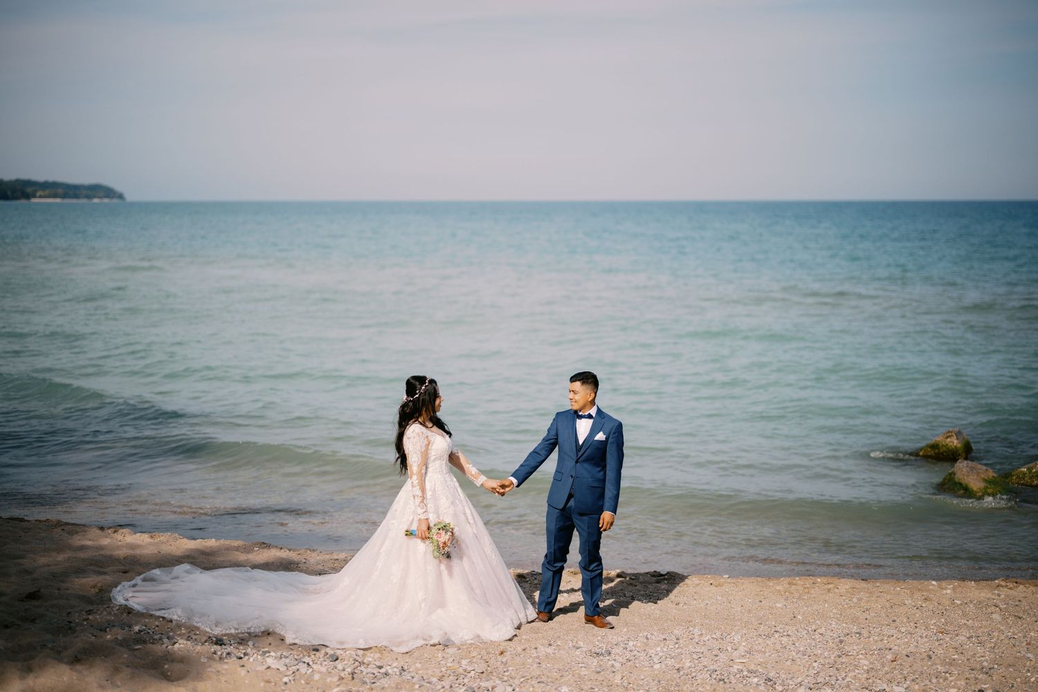 A couple in wedding attire holds hands on a serene beach with blue ocean waters stretching to the horizon.