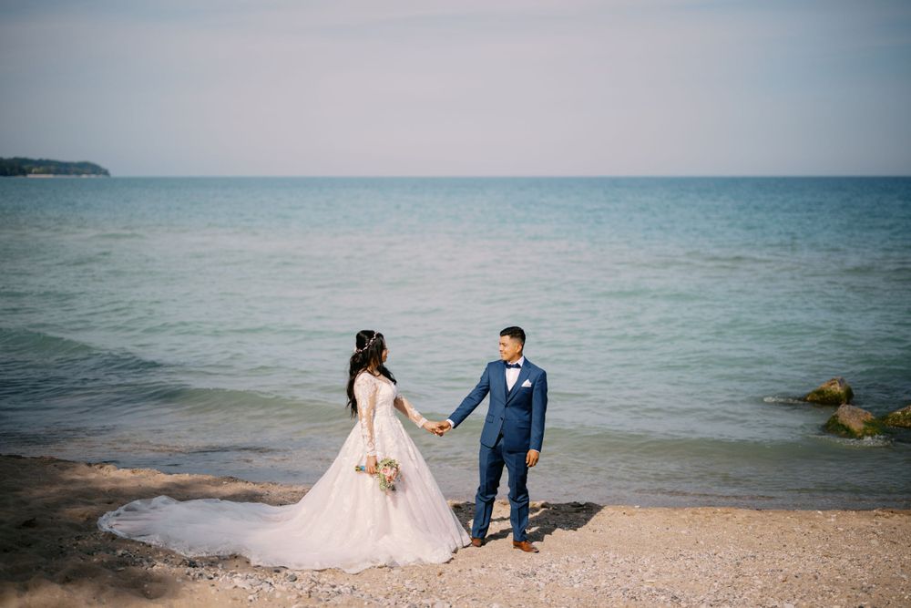 A couple in wedding attire holds hands on a serene beach with blue ocean waters stretching to the horizon.