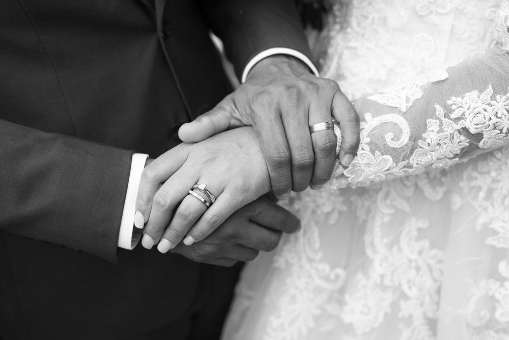 Close-up black and white photo of newlywed couple's hands showing wedding rings and lace dress details.