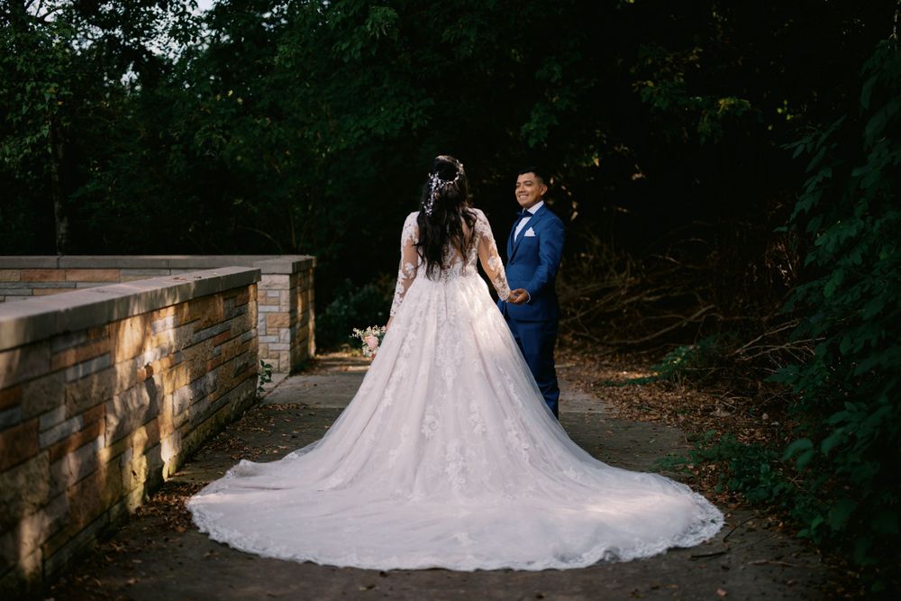 A couple in traditional wedding attire stands on a stone bridge path surrounded by lush greenery at sunset.