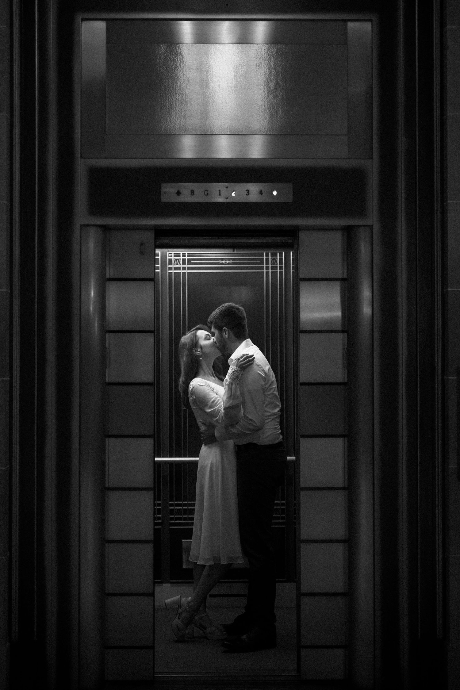 A romantic vintage-style black and white elevator scene with two figures embracing in an art deco interior.