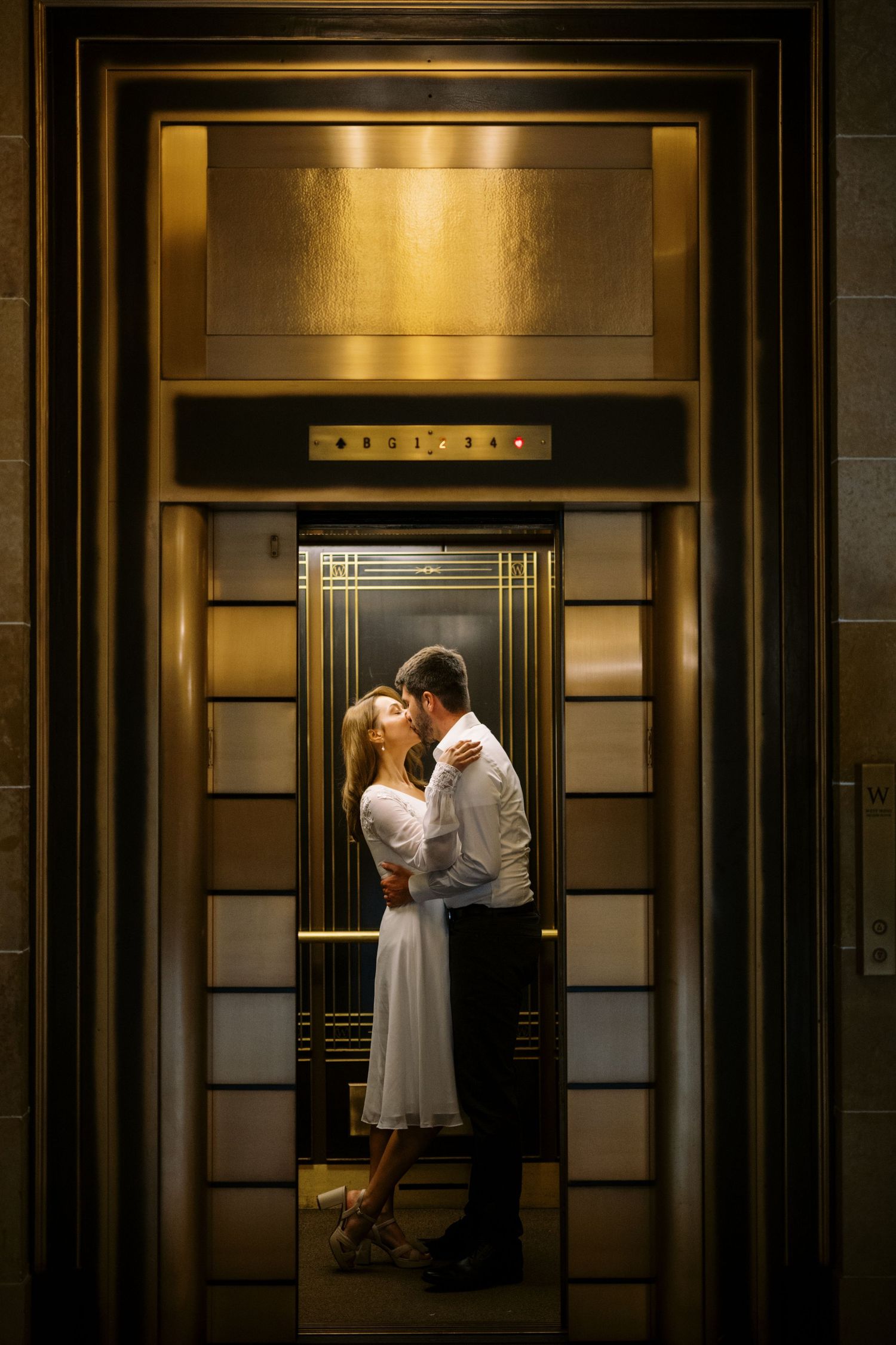 A romantic couple sharing an intimate moment in a warmly lit art deco style elevator with gold accents.