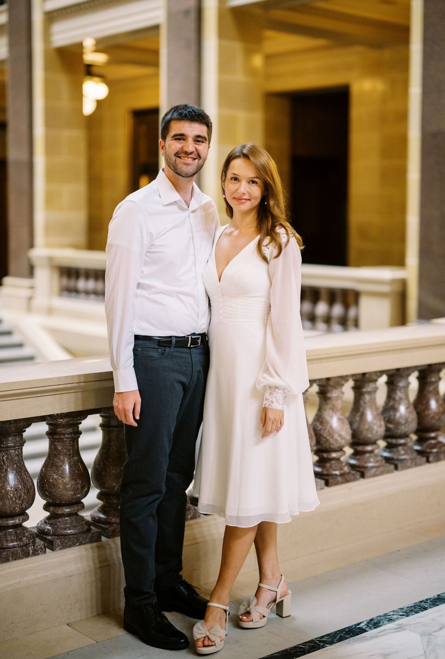 A well-dressed couple poses together on an elegant marble balcony in formal white attire.