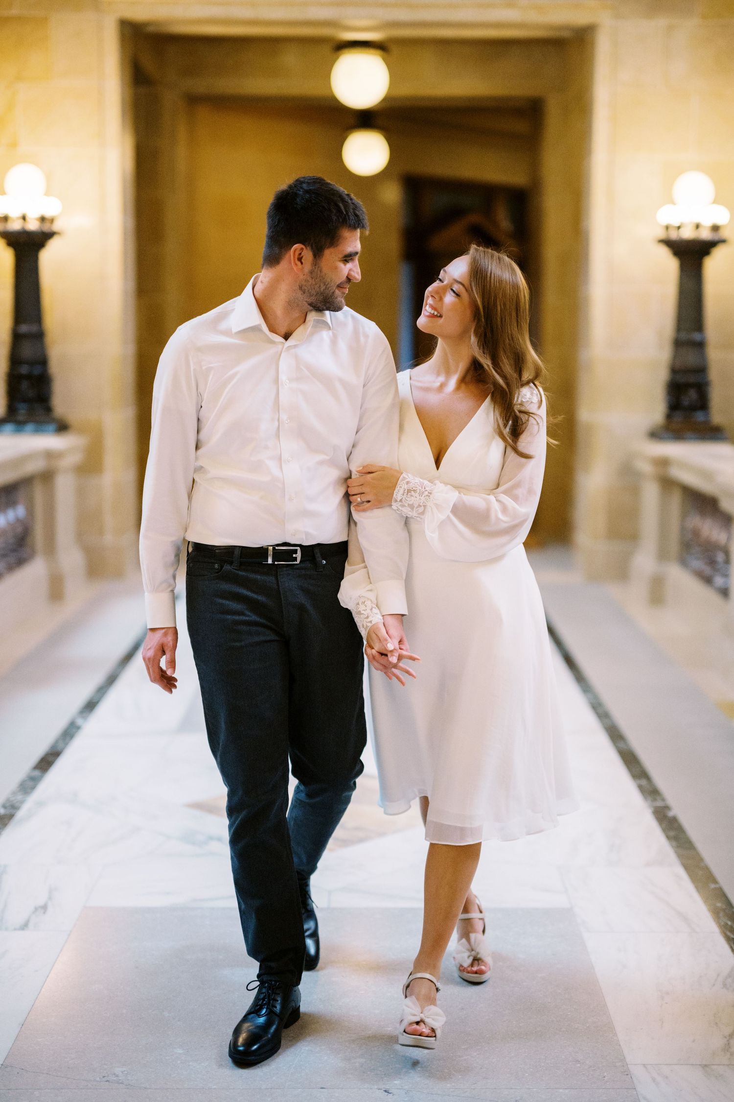 Couple walking down an elegant hallway with white marble floors and classic wall sconces.