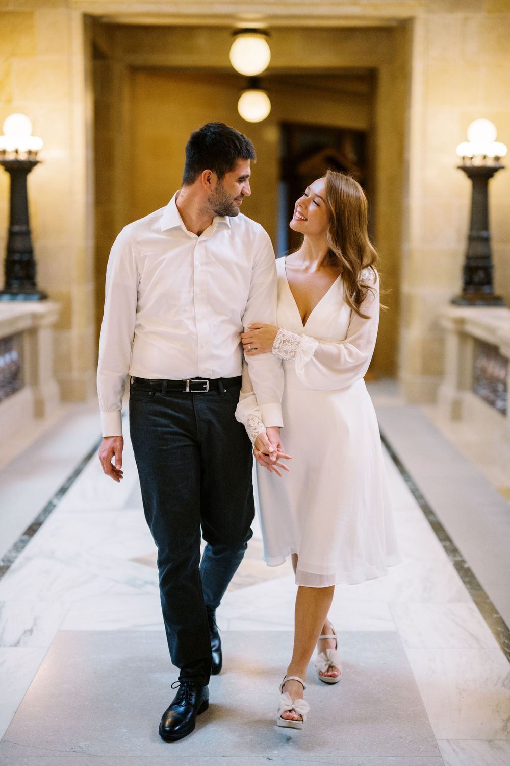 Couple walking down an elegant hallway with white marble floors and classic wall sconces.