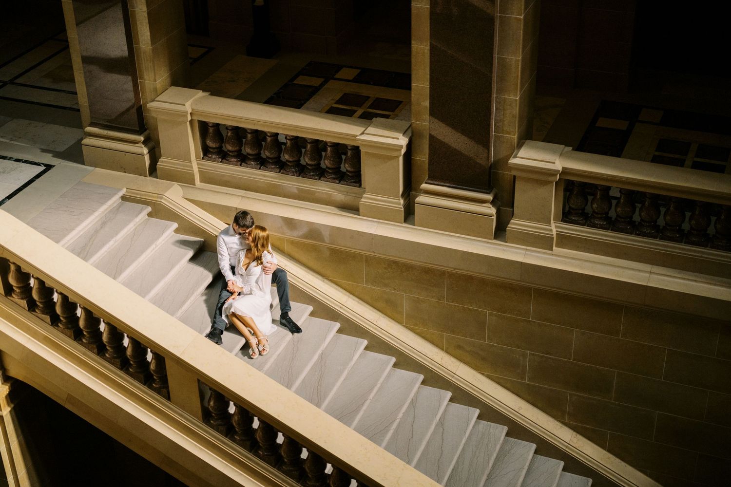 A solitary figure in white descends an ornate stone staircase illuminated by warm sunlight streaming in.
