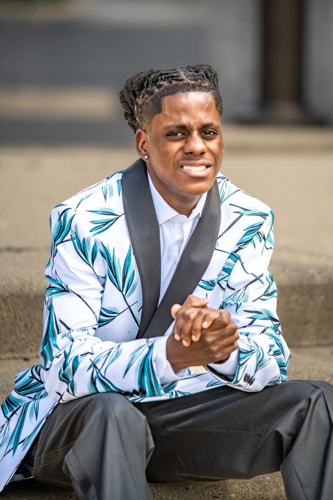 A person in a white and turquoise patterned blazer sits smiling on concrete steps.