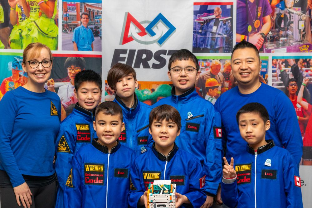 A FIRST robotics team wearing blue team uniforms poses together with their robot and competition display backdrop.