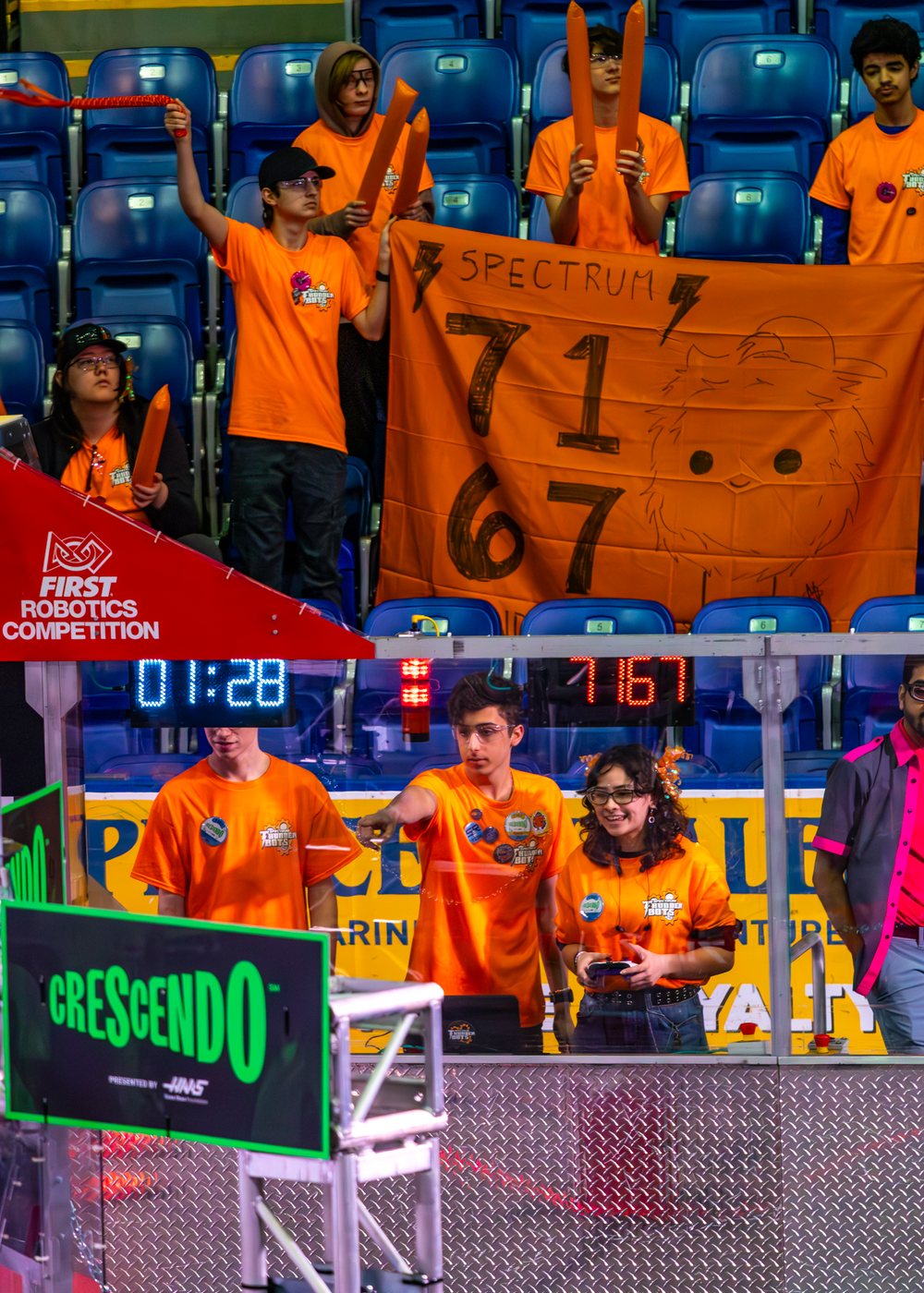 Teams holding orange banners in stadium seating during robotics competition with timer display visible.