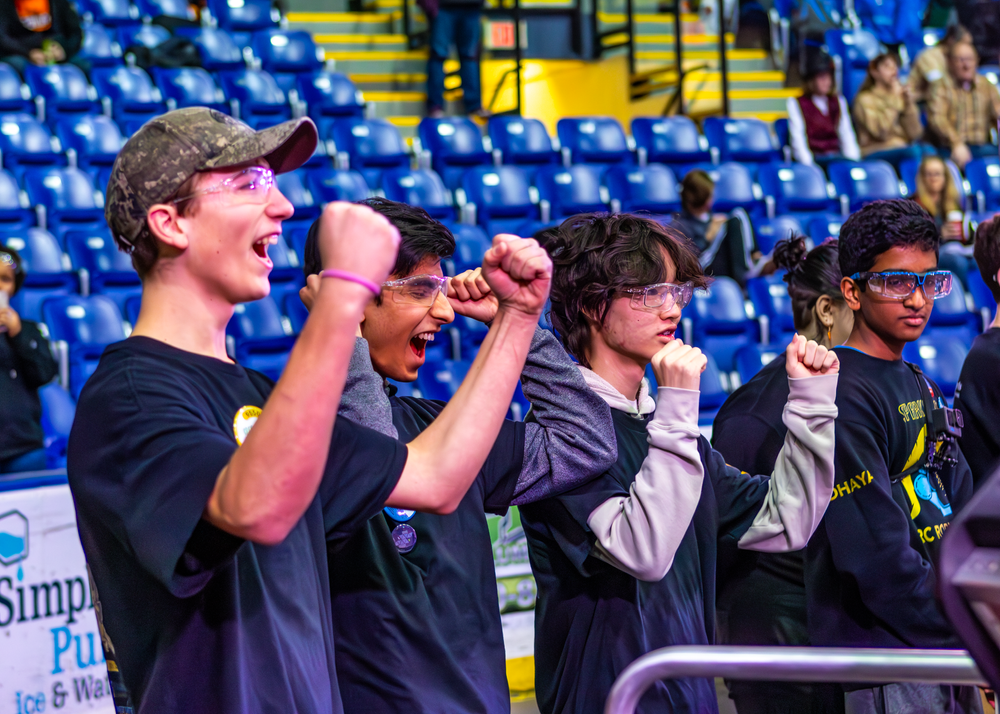 Competitors on stage celebrate during an esports tournament with blue stadium seating visible in background.