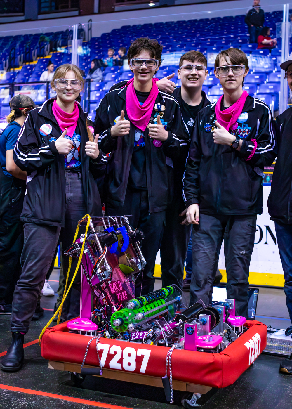 A competitive robotics team shows off their robot build on a red platform at a competition venue with blue stadium seating.