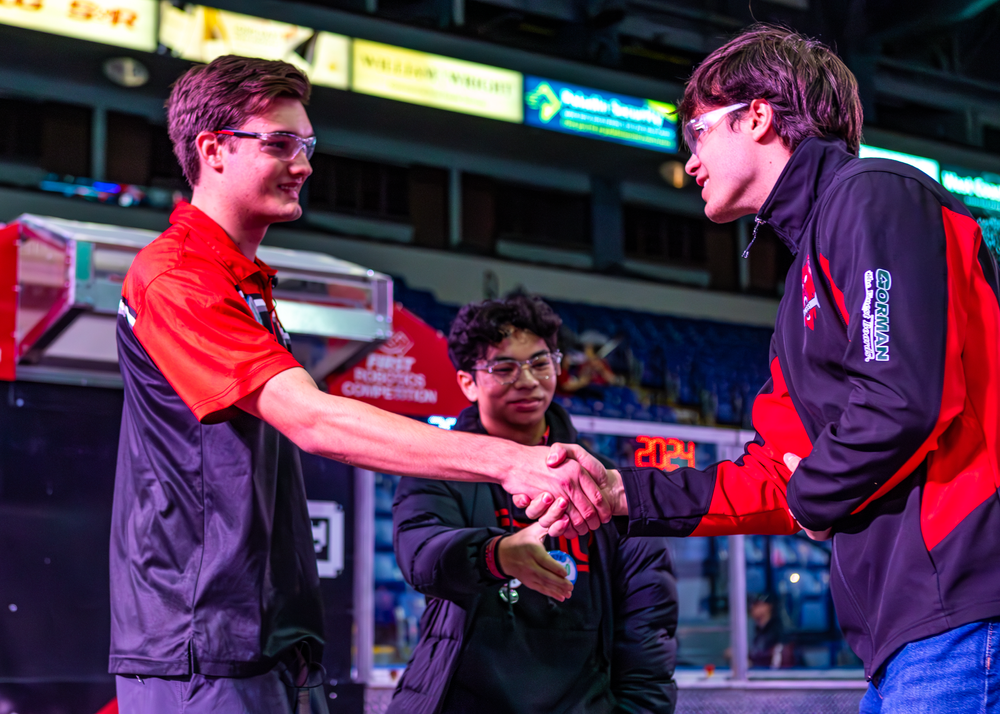 Competitors in a robotics competition shake hands in a display of sportsmanship under colorful lighting.