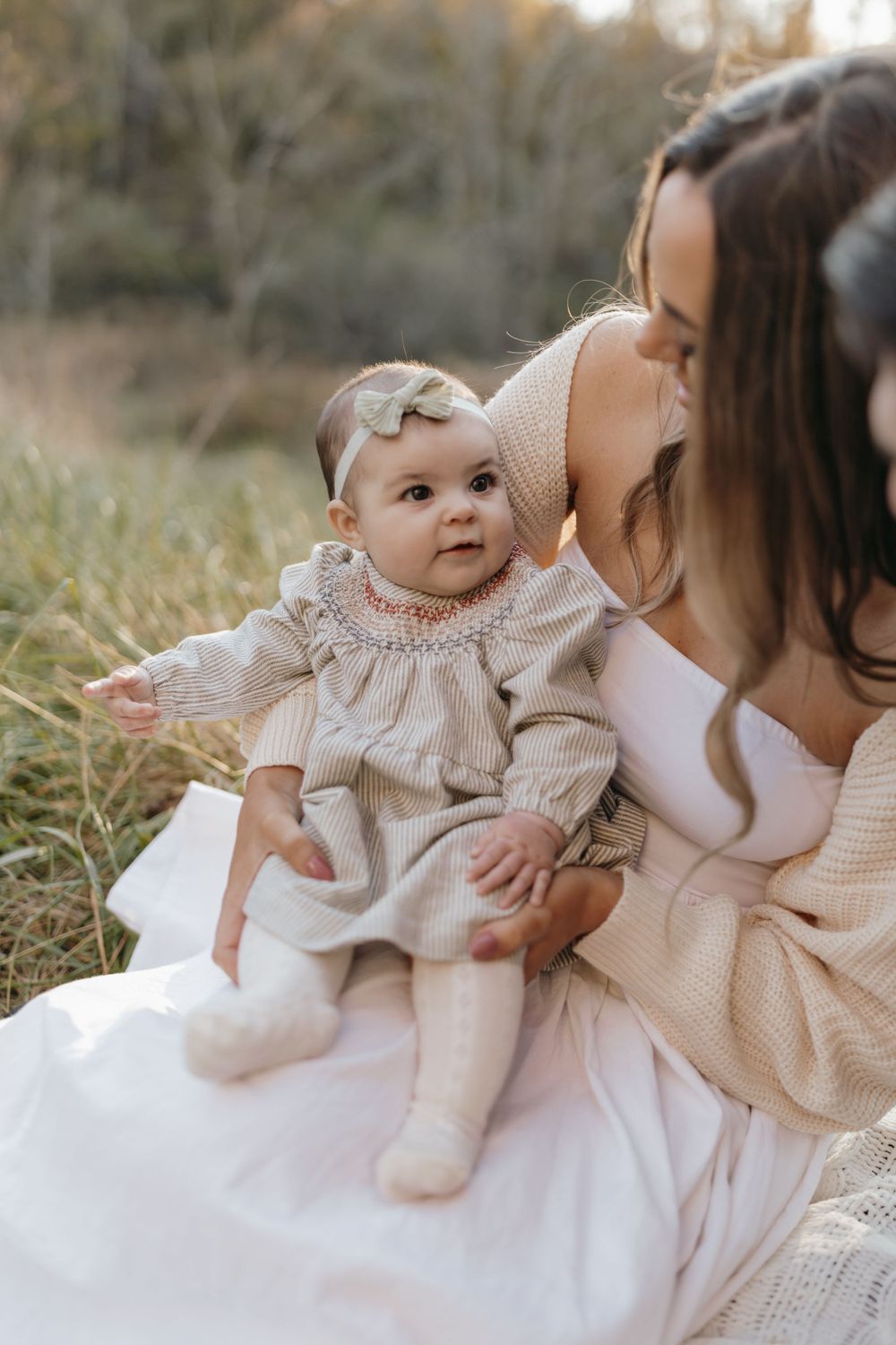 A baby in a delicate smocked dress sits on a white blanket outdoors during a golden hour photoshoot.