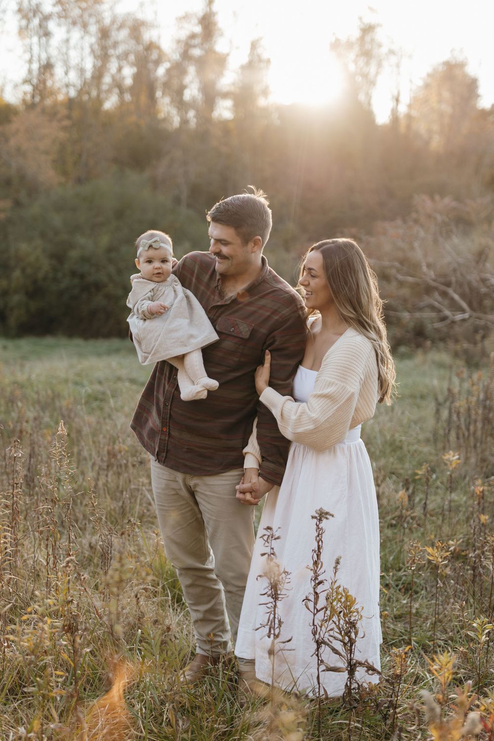 A family stands together in a sunlit meadow during golden hour wearing coordinated neutral outfits.