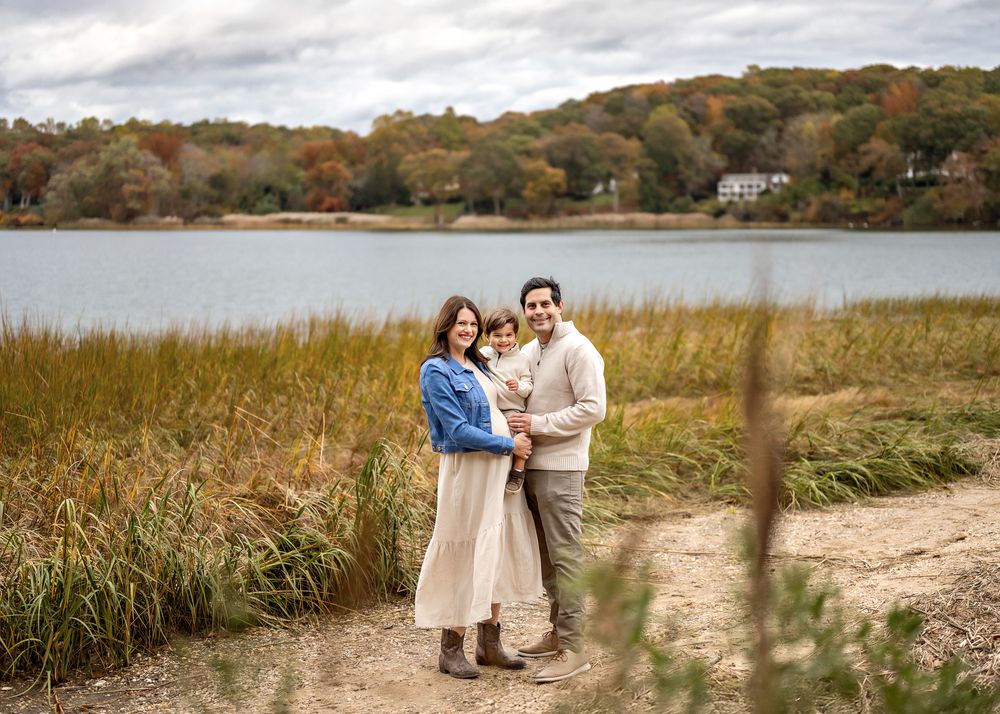 A couple stands together on a lakeside path surrounded by autumn foliage and tall grass on a cloudy day.