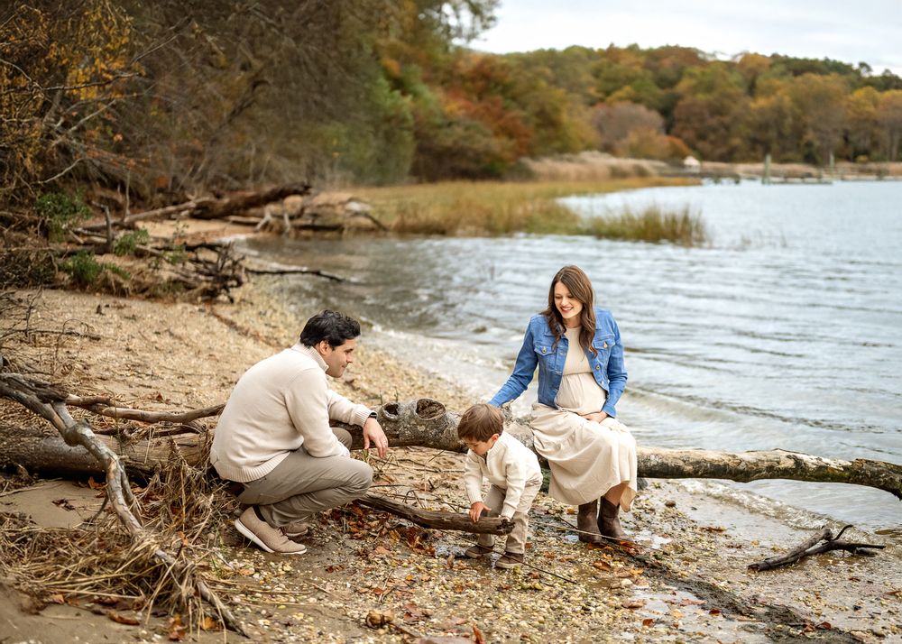 A couple sits together with their dog on a rocky lakeshore during autumn with trees in the background.