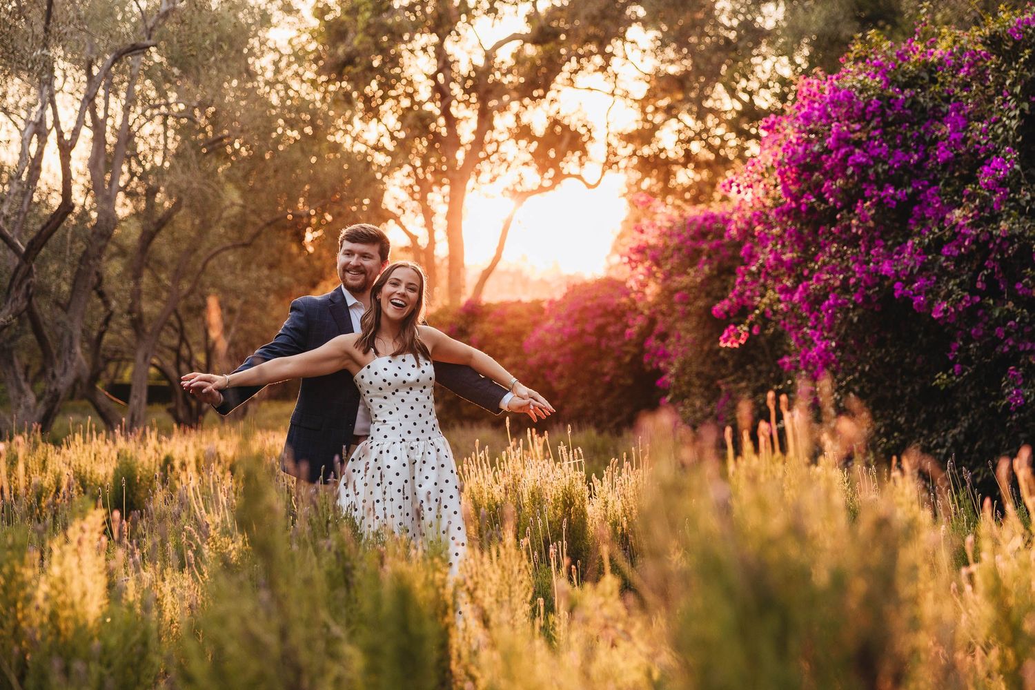 Couple holding hands and laughing together on green lawn with autumn foliage in background.