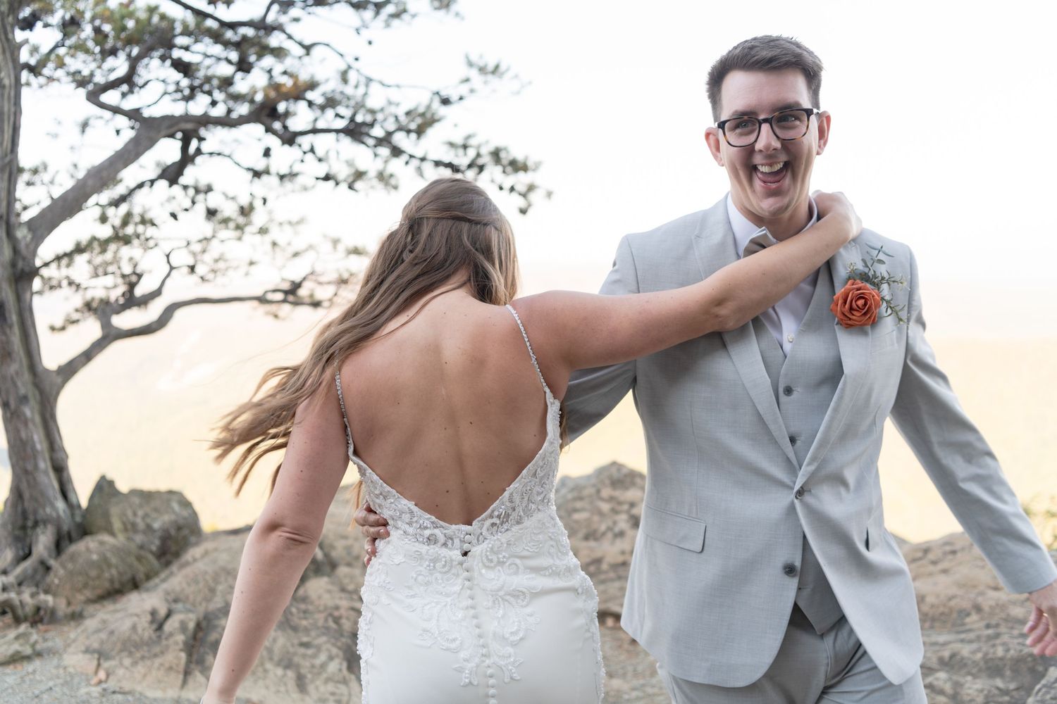 Wedding couple share a joyful moment together on a mountaintop at sunset wearing formal attire.