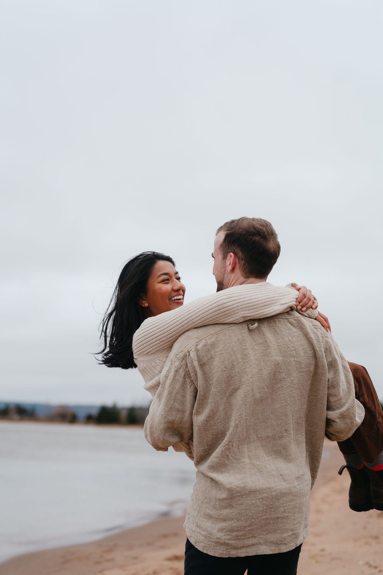 A couple embraces on a beach with overcast skies and water in the background.