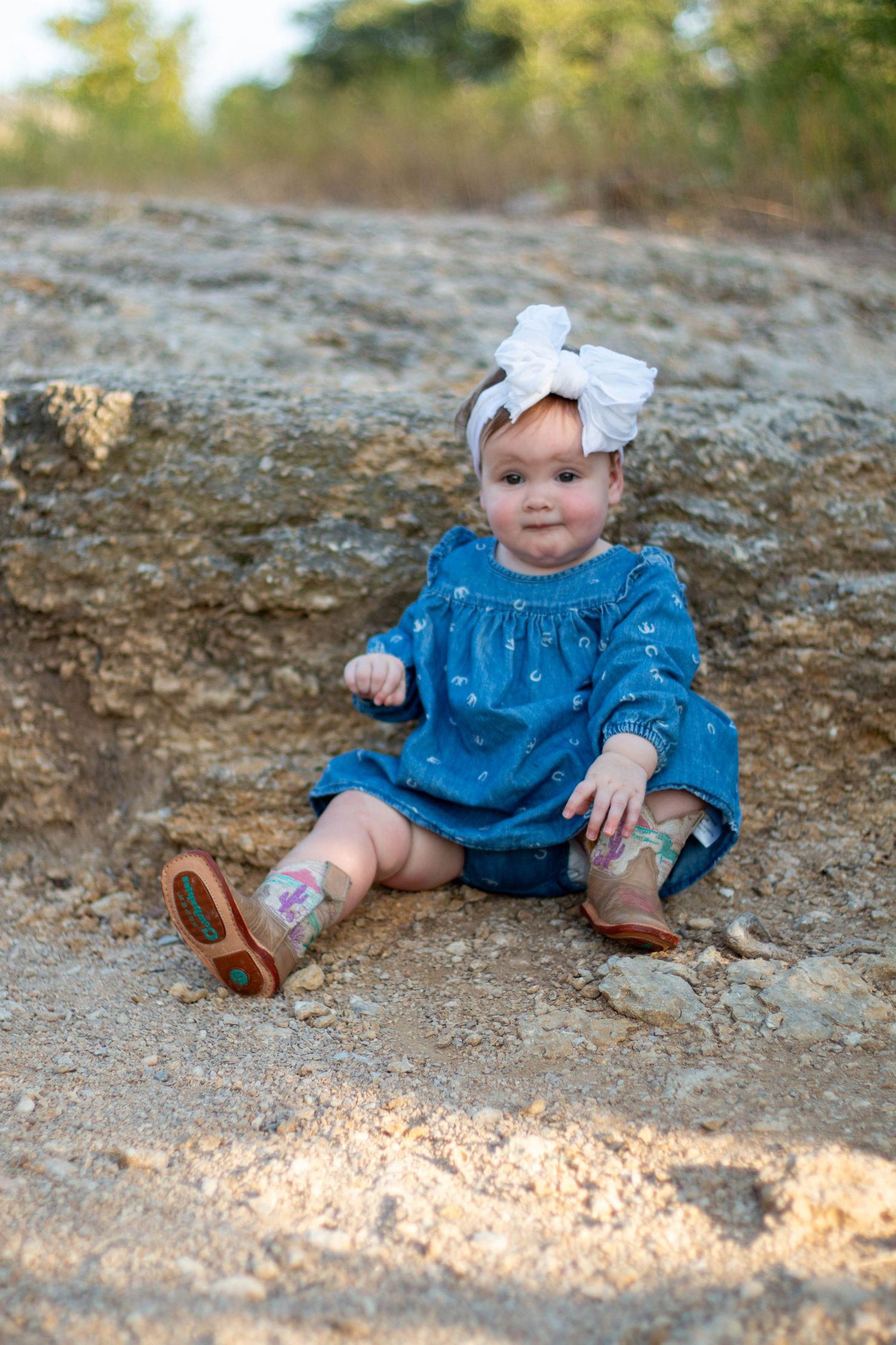 Happy baby in blue overalls sits on white blanket playing with colorful plastic Easter eggs scattered around them.