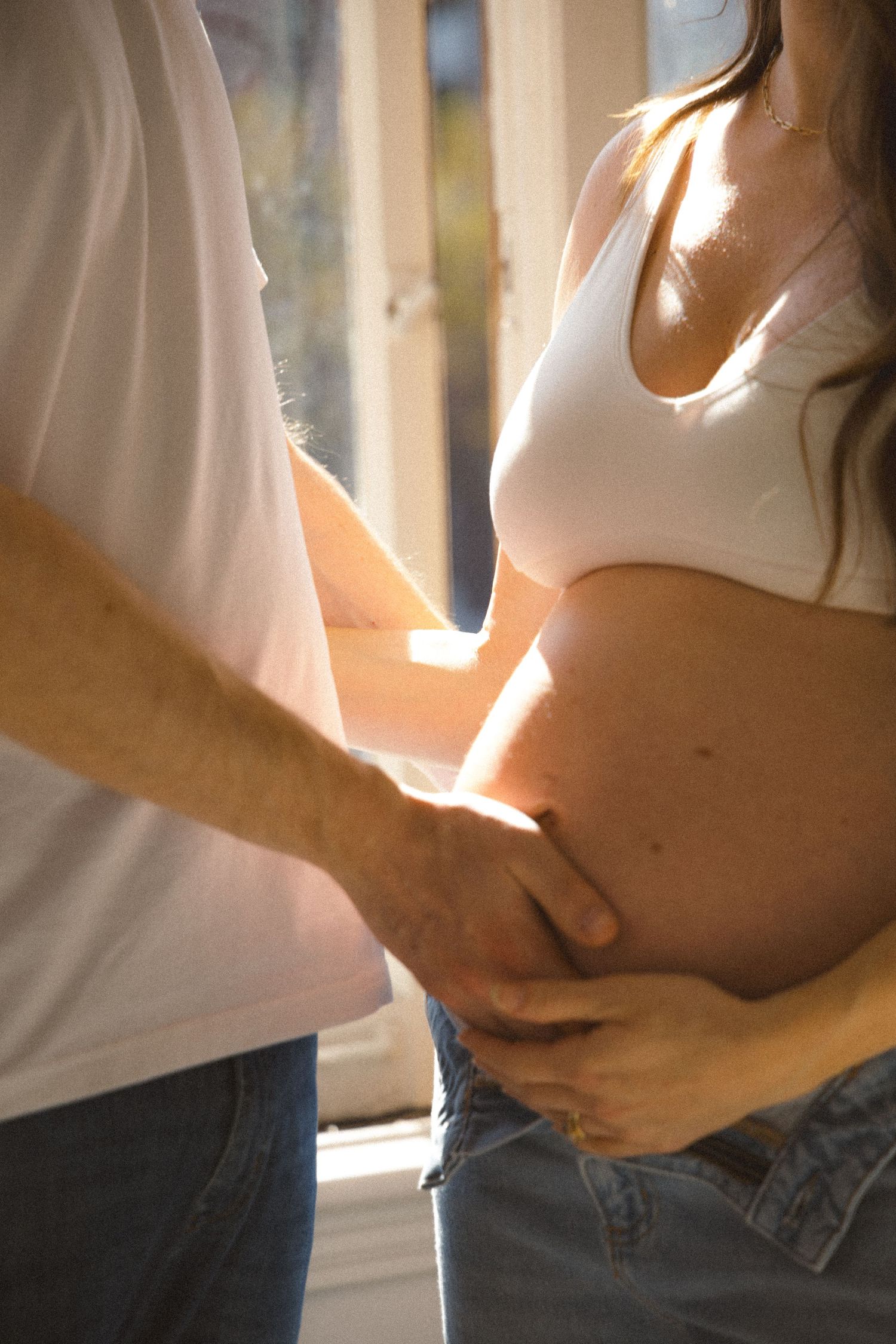 Couple dressed in white looking out of the window in natural sunlight during a family maternity photosession in a studio in Vancouver, BC.