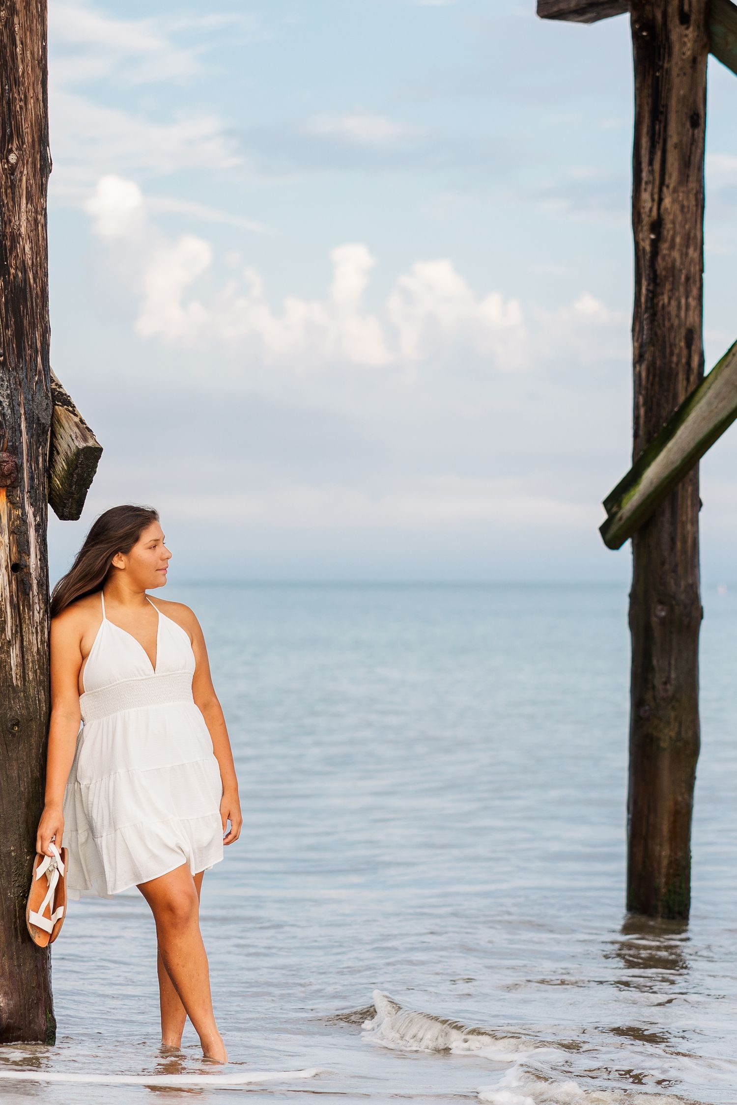 Person in a flowing white dress walking along the beach shoreline near wooden pier posts at sunset.