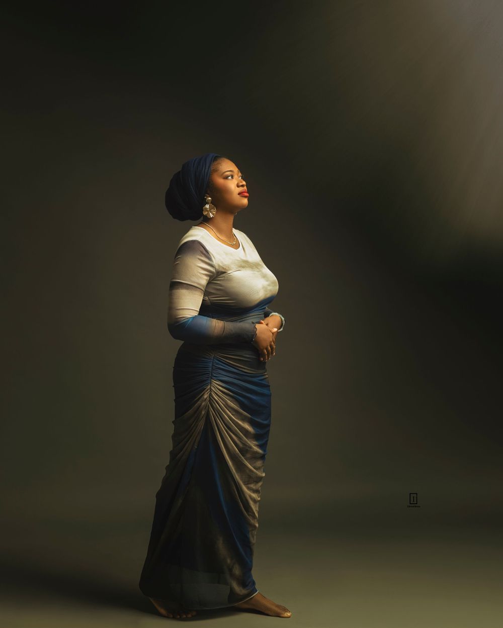 Dramatic studio portrait in fitted top and draped skirt with head wrap against dark background.