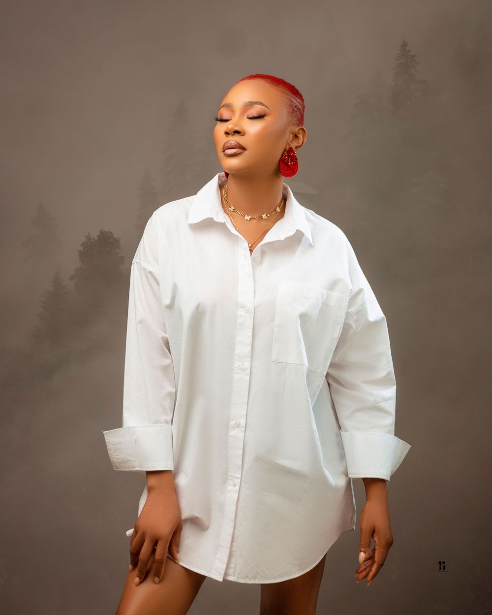 Model in oversized white button-down shirt dress with short red hair poses against moody outdoor background.