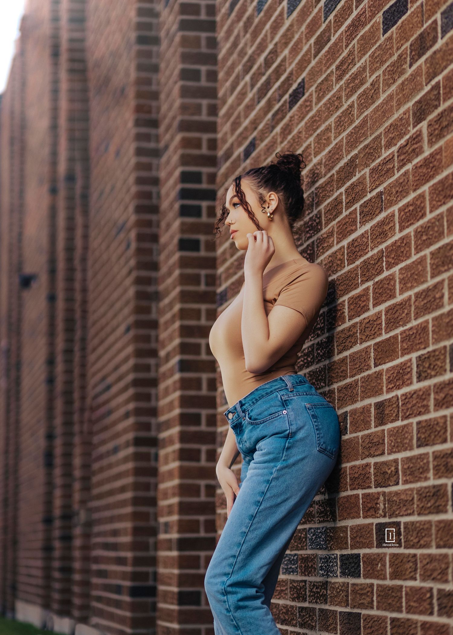 Woman in beige top and high-waisted jeans stands against rustic brick wall in urban setting.