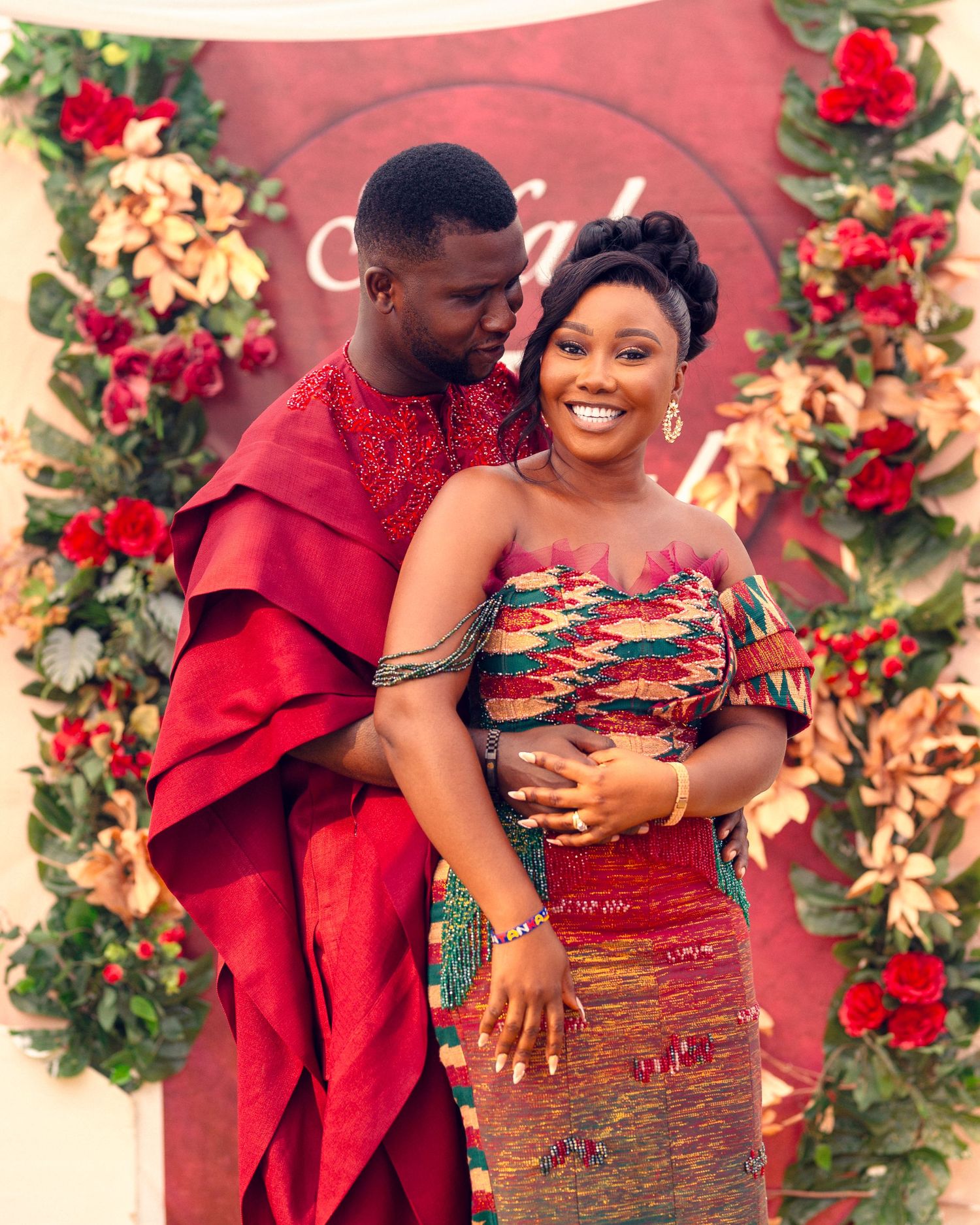 Couple in traditional African dress share tender moment against backdrop of red roses and decorative flowers.