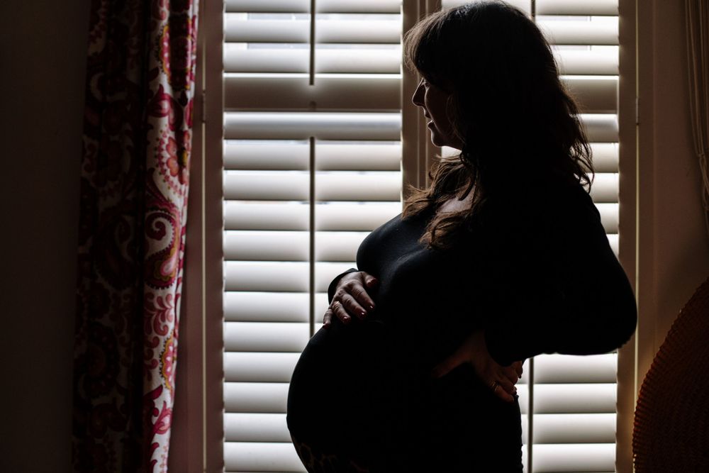 A silhouetted pregnant figure stands by a window with plantation shutters, tenderly holding her baby bump.