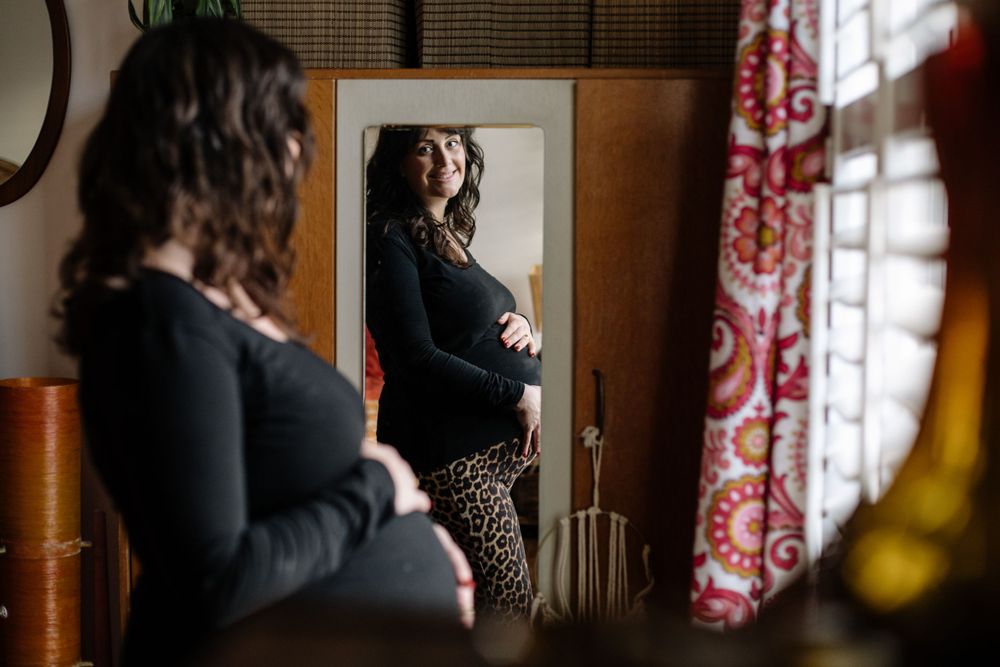 A pregnant figure is reflected in a mirror next to red curtains in a warmly lit room during a maternity photoshoot.