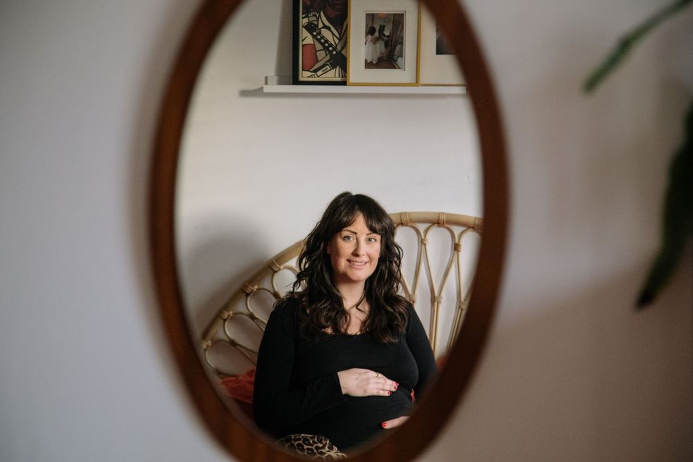 A mirror reflection shows someone in a black sweater sitting in a rattan chair against a white wall with framed artwork.