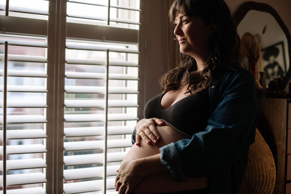 An expectant mother stands near window blinds in moody lighting with a casual sweater draped around her.
