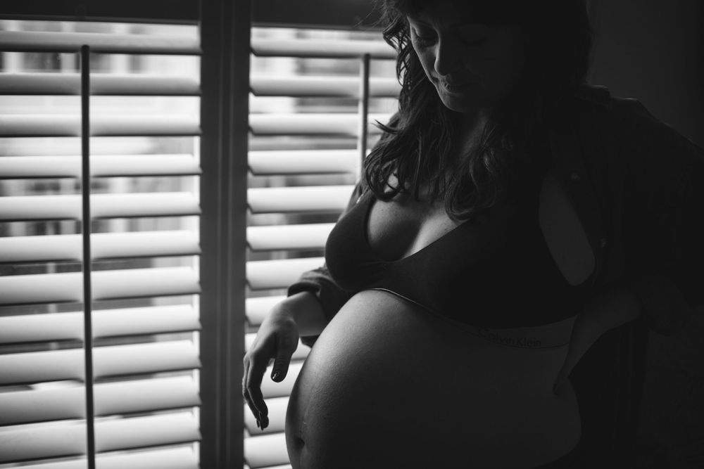 Silhouette maternity photo with light filtering through window blinds creating dramatic shadows.