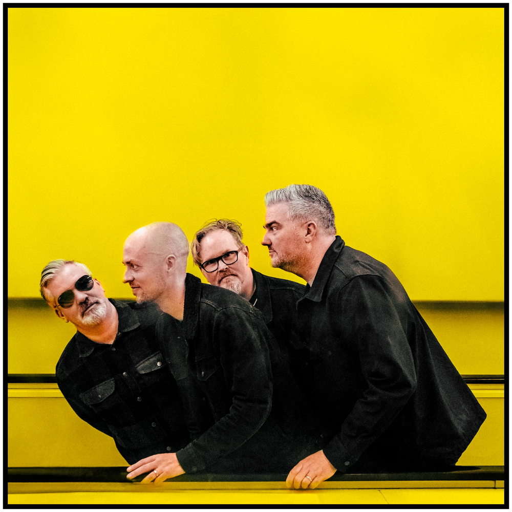 Four band members posing playfully against bright yellow background wearing dark clothing. Portland music photographer
