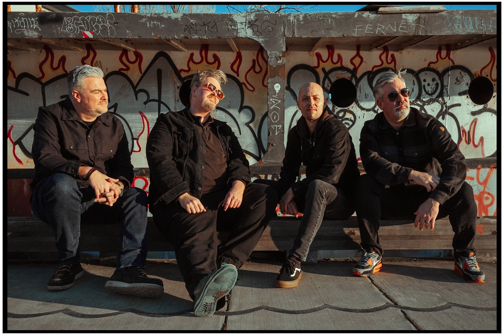 Four band members in dark clothing sitting against graffiti-covered wall. Portland music photographer