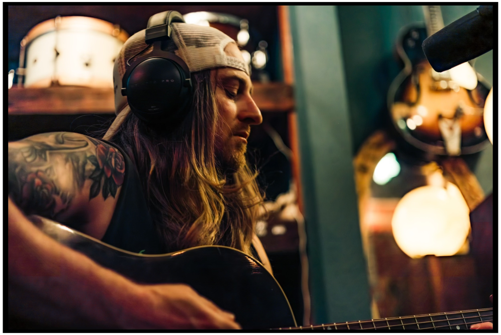 Musician with long hair and tattoos playing guitar in dimly lit studio setting. Portland music photographer