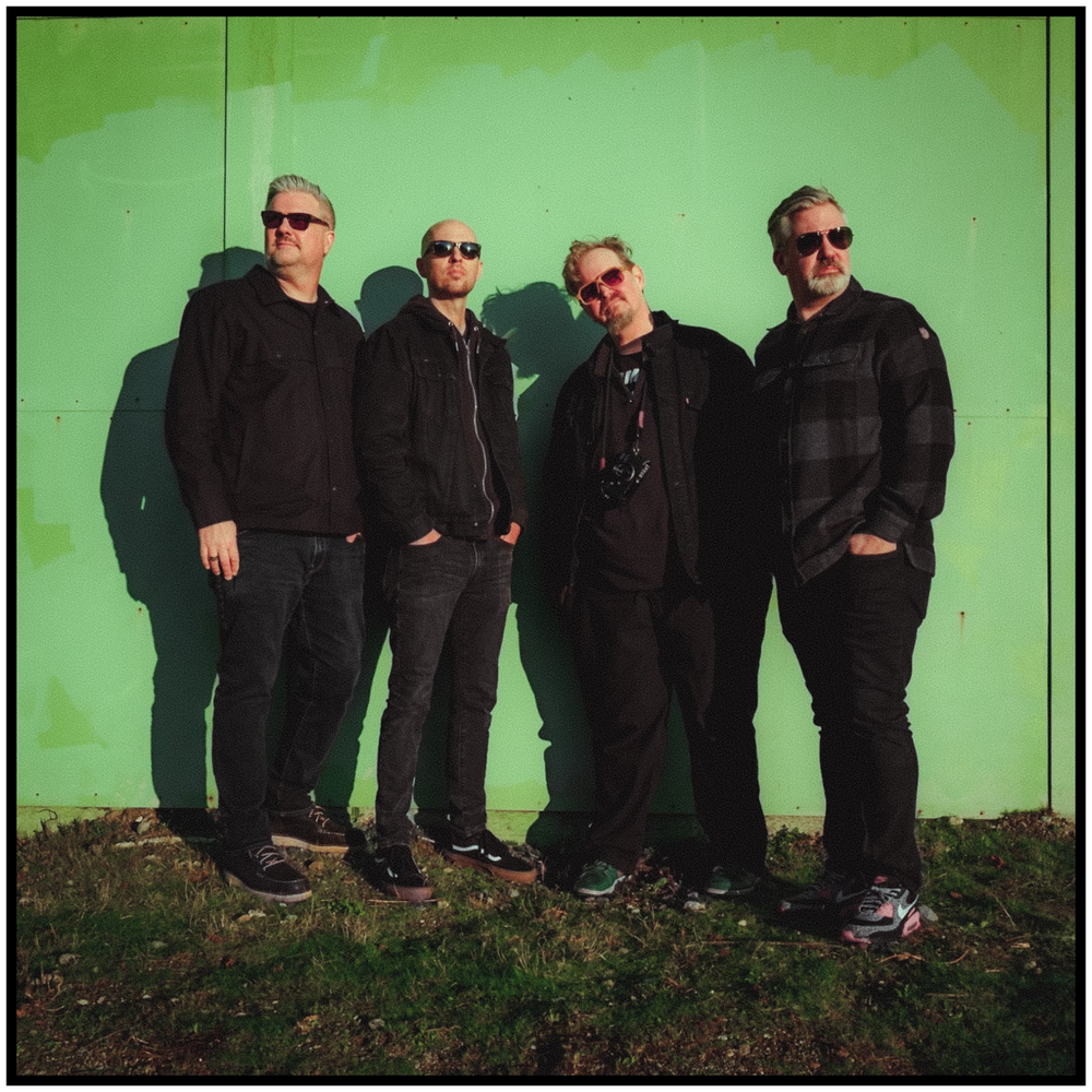 Musical group in dark clothing poses against green wall for promotional photo. Portland music photographer
