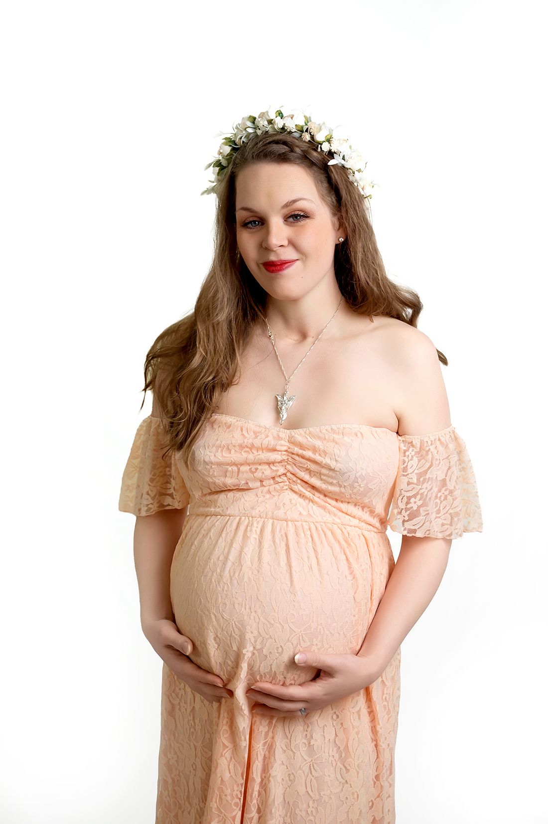 A pregnant woman in a peach-colored off-shoulder lace dress wears a delicate flower crown against a white backdrop.