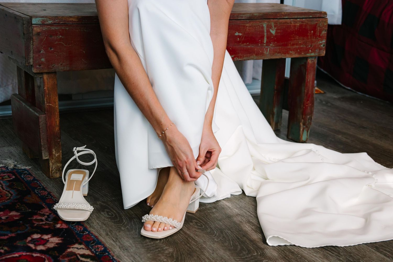 Someone in a white wedding dress adjusts elegant sandal straps while sitting on a rustic wooden bench.