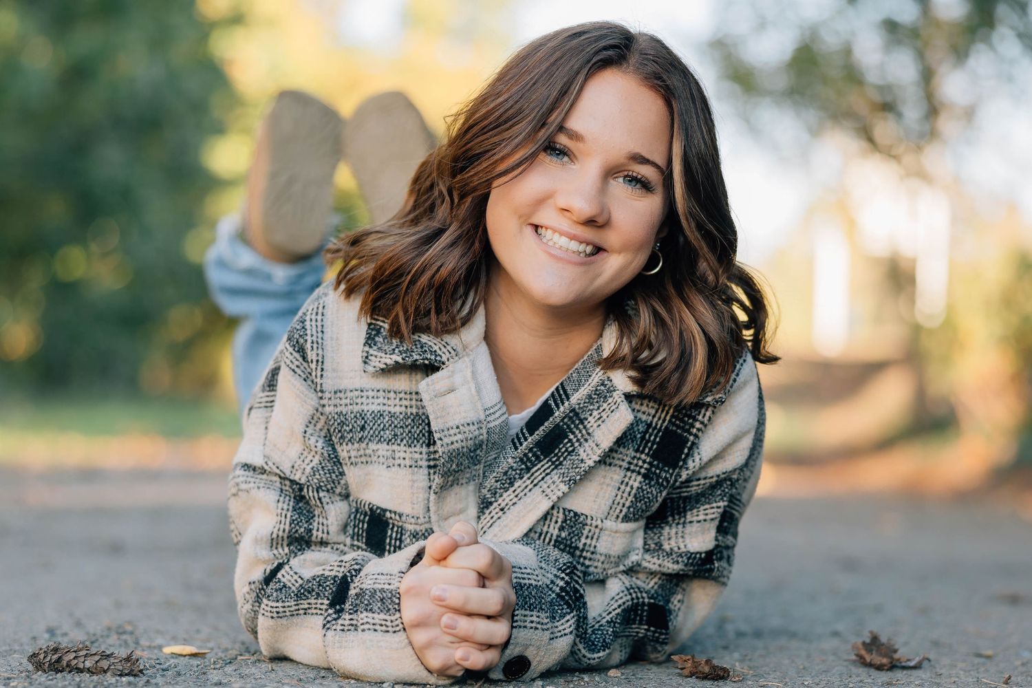Smiling teenage girl in plaid jacket lying on the ground outdoors in a fall setting.