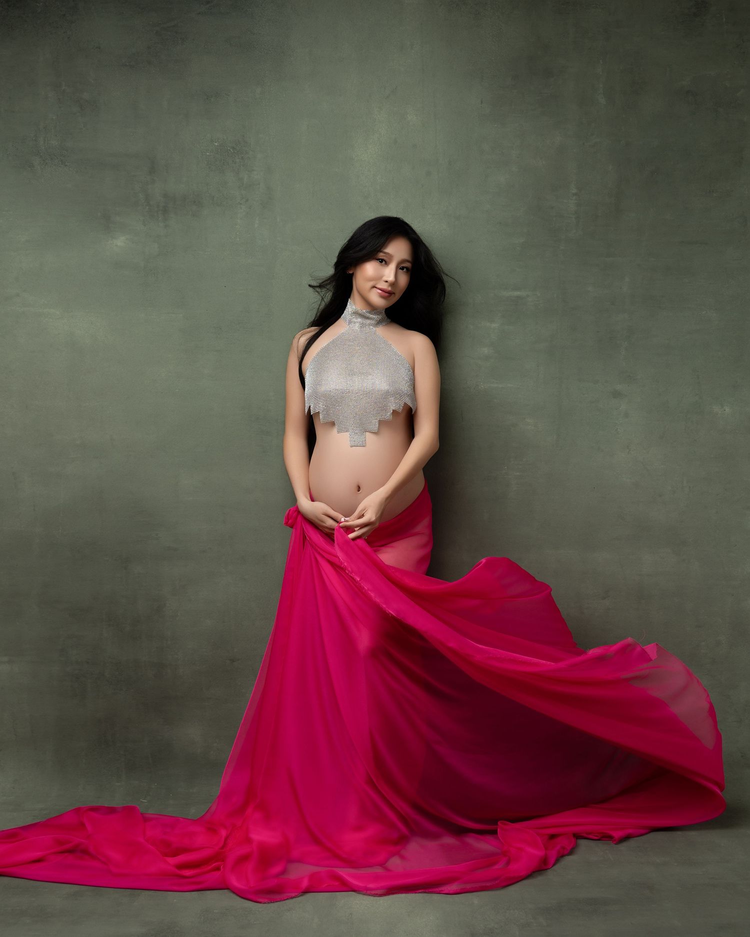 A figure poses in a silver sequined crop top and flowing magenta skirt against a textured green background.
