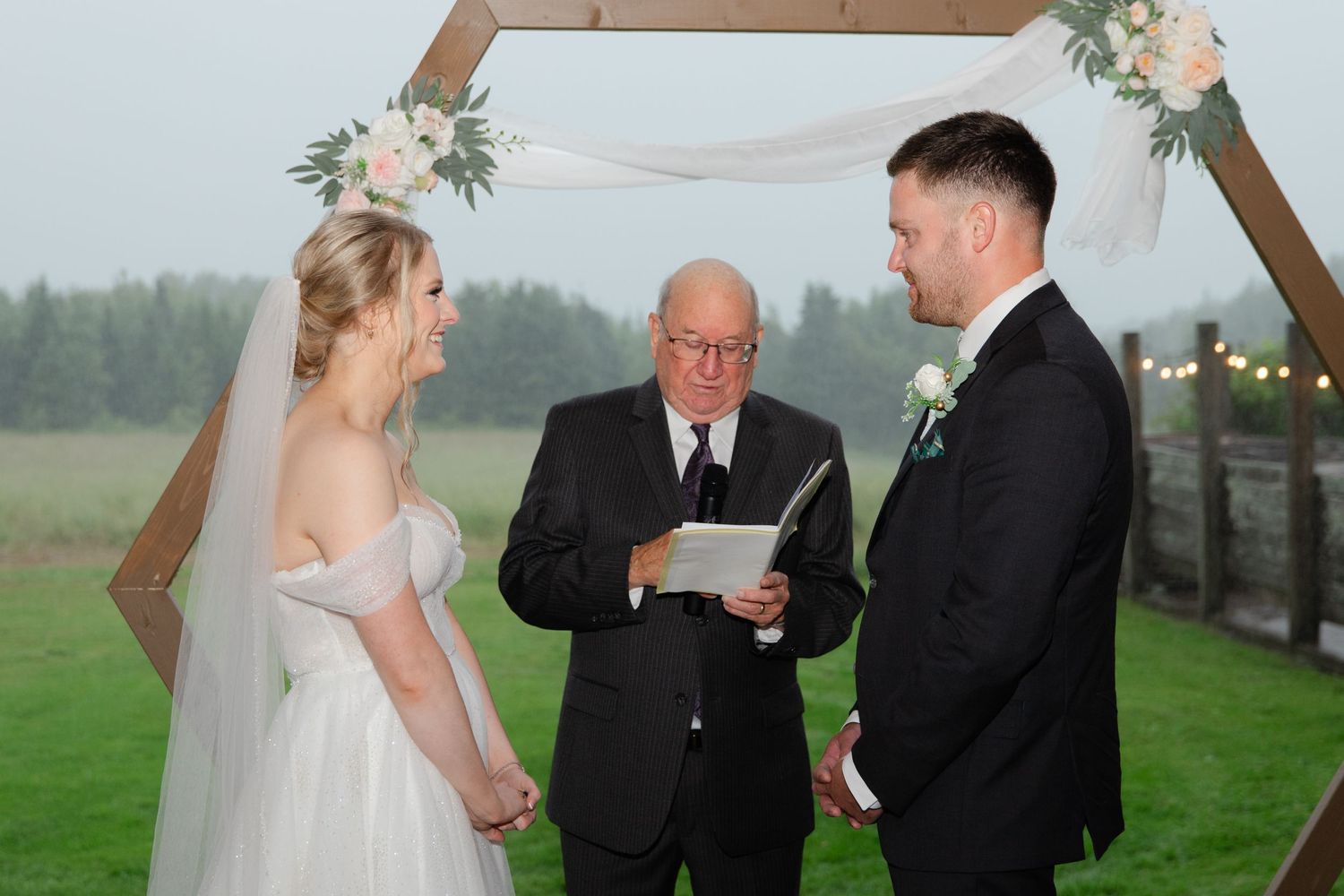A wedding ceremony takes place outdoors beneath a wooden arch decorated with white flowers as the officiant reads from papers.