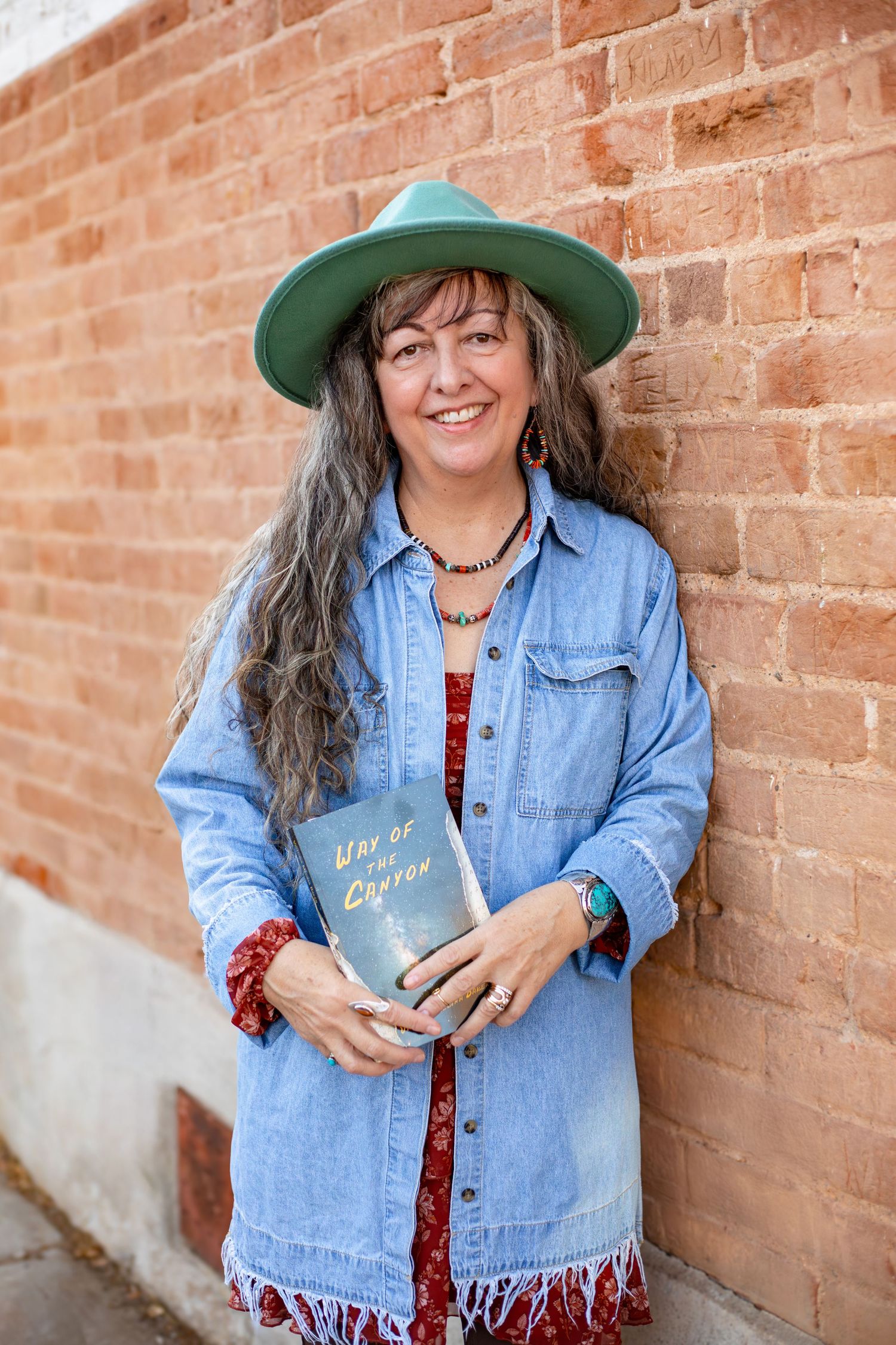 Lifestyle portrait against red brick wall wearing denim jacket and holding a book.