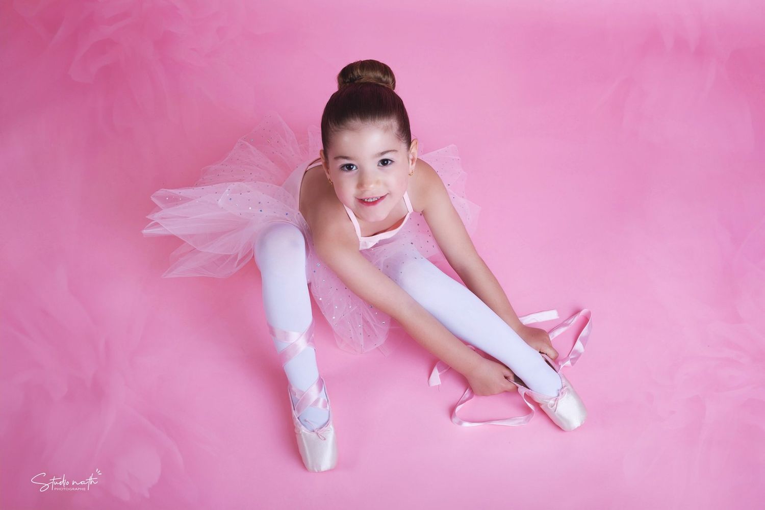 A young ballet dancer in white tutu and pointe shoes poses gracefully against a pink background in a studio setting.