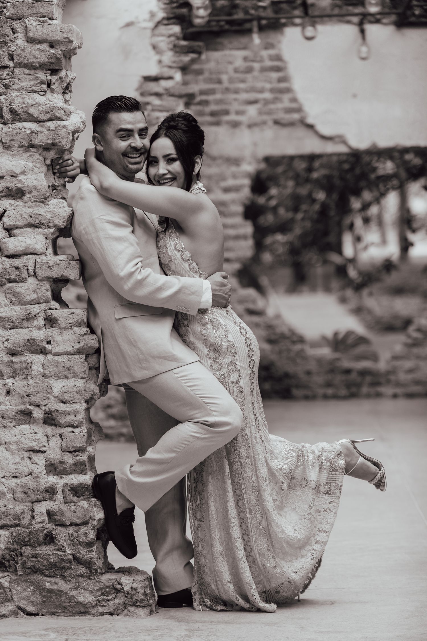 A romantic black and white portrait of a couple embracing against a brick wall, the woman wearing a sparkly gown.